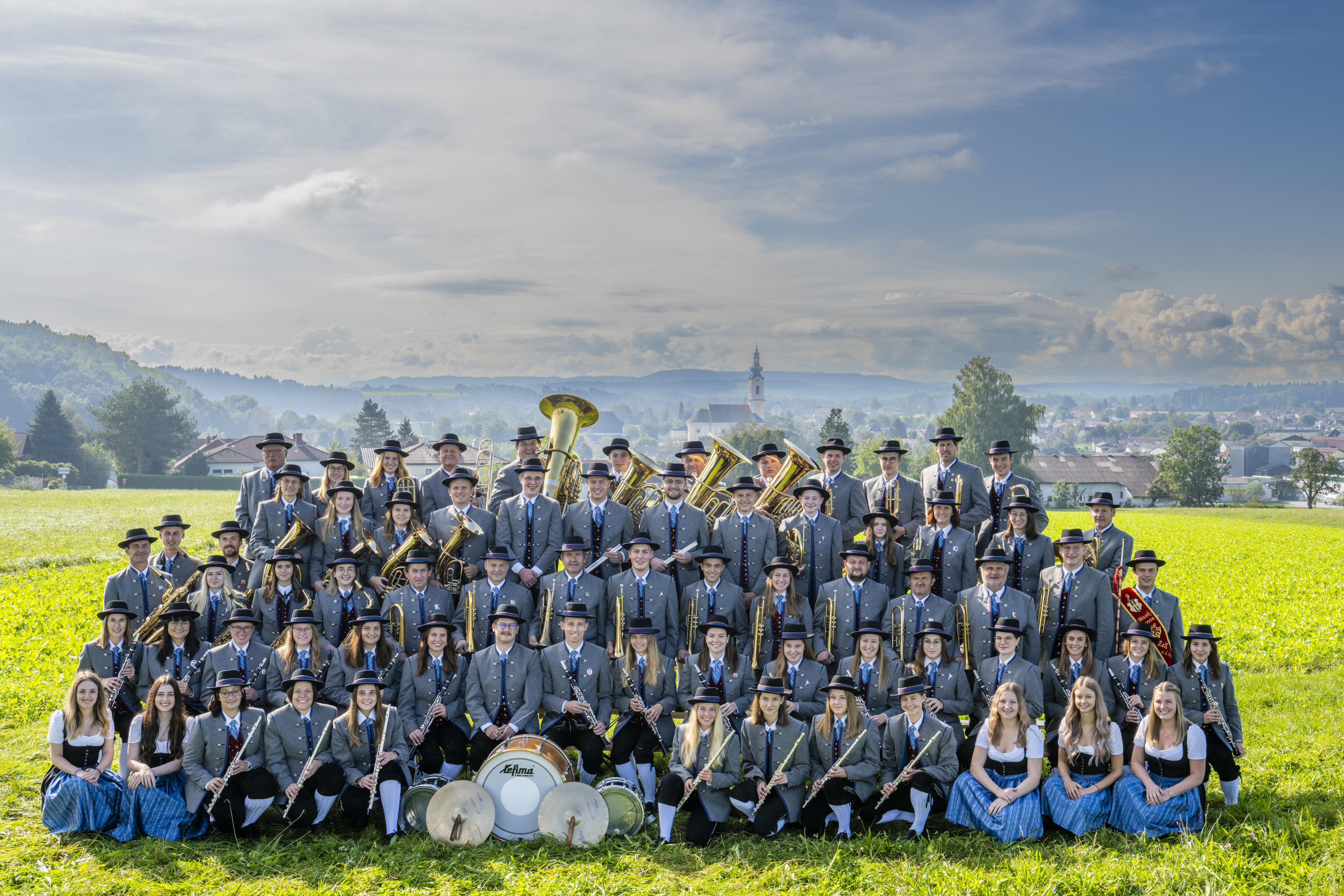 Eine Gruppe von ca. 60 Musikanten einer Musikkapelle auf einer grünen Wiese, auf einem Gruppenfoto in sechs Reihen angeordnet, alle in Tracht gekleidet, mit verschiedenen Musikinstrumenten einer Blasmusikkapelle in der Hand, im Hintergrund blauer Himmel mit Wolken, hinter den Musikern sin der Ferne ein Dorf mit Häusern und einer Kirche genau in der Bildmitte